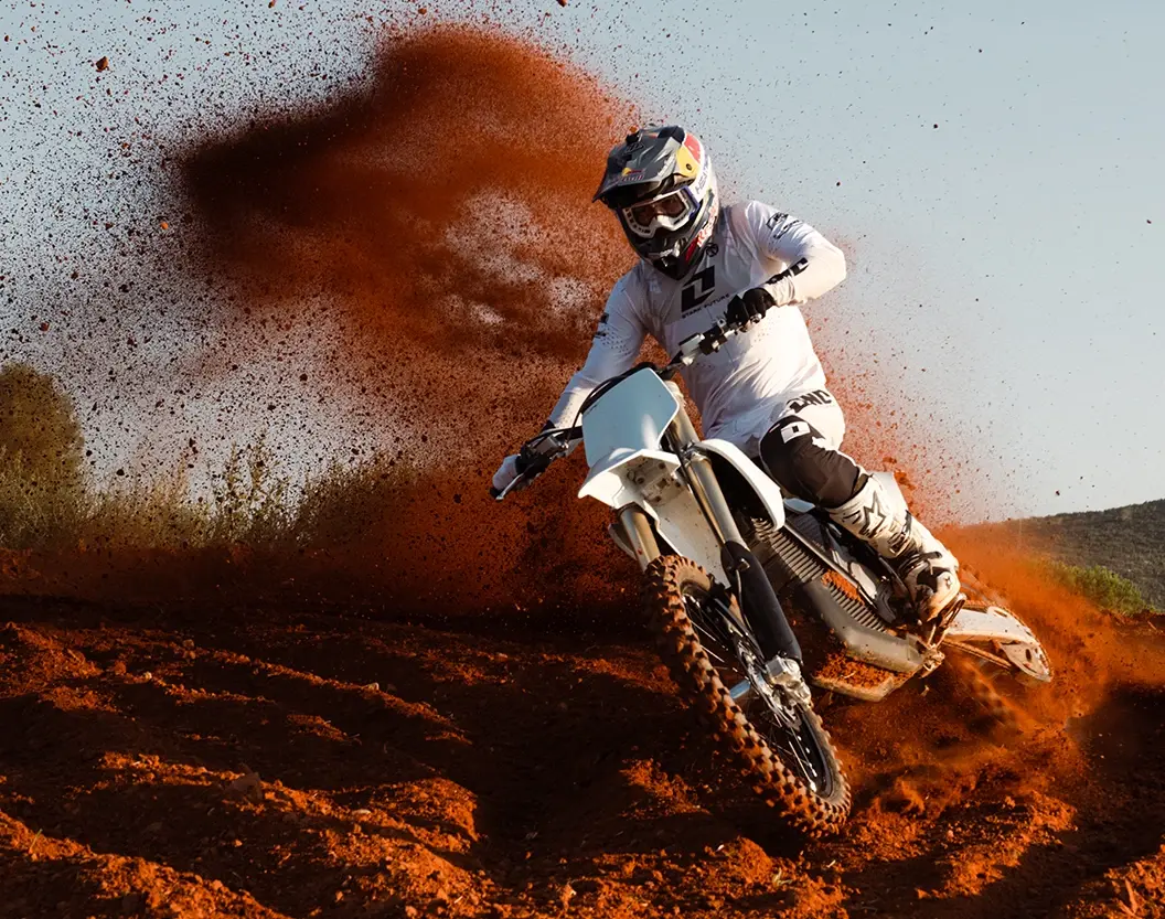 Rider cornering a white Stark Varg hard on a dry orange motocross track, throwing a roost of dirt